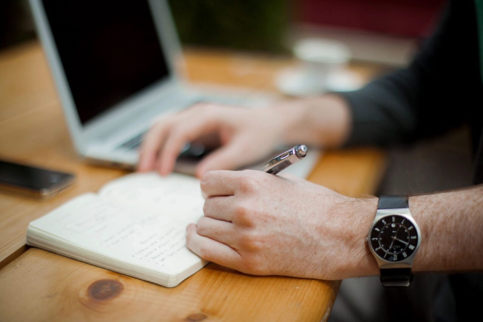 Person writing notes near an open laptop.