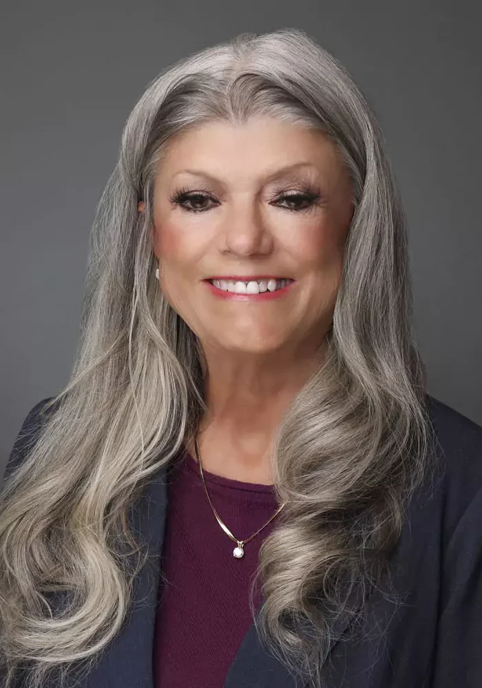 Smiling woman with long gray hair wearing a dark blazer and maroon top.